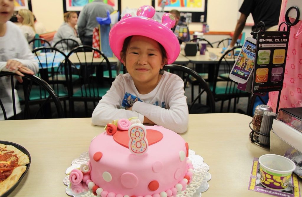 Girl with fun balloon birthday hat on sitting in front of a polka-dot pink birthday cake.